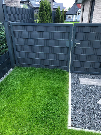 Ein moderner grauer Zaun mit einem passenden Tor trennt einen üppigen grünen Rasen von einem Kiesweg in einem Wohnhinterhof. Im Hintergrund sind Bäume und Häuser zu sehen.