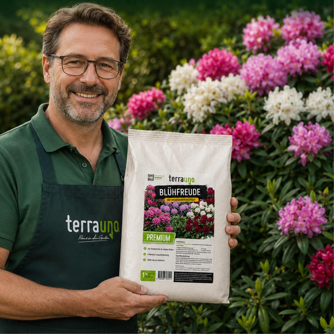 Ein lächelnder Mann mit Brille und einer grünen Terrauno-Schürze hält eine Tüte „Blühfreude Premium“-Pflanzendünger in der Hand, im Hintergrund ist ein Garten mit blühenden rosa und violetten Blumen zu sehen.