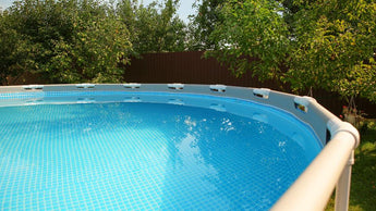 Ein oberirdischer Swimmingpool mit klarem, blauem Wasser liegt in einem Hinterhof, umgeben von grünen Bäumen und einem braunen Holzzaun an einem sonnigen Tag.