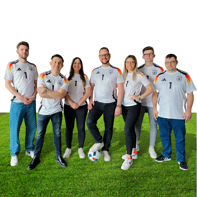 Sieben Personen stehen auf dem Rasen und tragen passende weiße Deutschland-Fußballtrikots. Eine Person hält einen Fußball mit dem Fuß fest. Sie lächeln, schauen in die Kamera und posieren gemeinsam vor einem weißen Hintergrund.