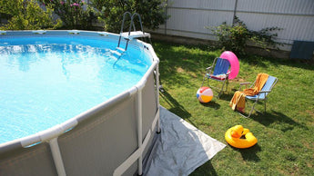 In einem sonnigen Hinterhof ist ein oberirdisches Schwimmbecken mit einer Leiter aufgestellt. In der Nähe stehen zwei Liegestühle, Handtücher, ein rosa Schwimmring, ein Wasserball und eine gelbe Ente, die auf grünem Gras schwimmt. Im Hintergrund sind Sträucher und ein Zaun zu sehen.