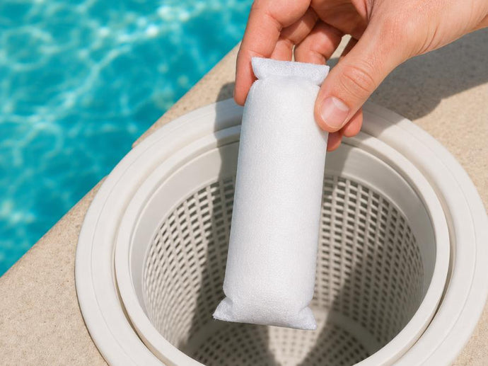 Eine Hand, die eine weiße Chlor-Tablette oder einen Desinfektionsmittelbeutel über den Skimmerkorb am Rand eines Schwimmbeckens hält.