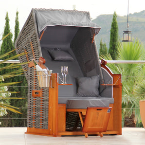 Der DERKORB Der Klassiker 1-Sitzer ist ein Loungesessel im Strandkorb-Stil aus Polyrattan und Kiefernholz, mit Baldachin, grauen Kissen, Fußstützen, Seitentablett mit Weingläsern und Eimer - perfekt zum Entspannen auf Ihrer Terrasse.