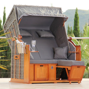 Der DERKORB „Der Klassiker 2-Sitzer“ vereint wetterbeständigen Komfort und maritimes Flair: großer Strandkorb aus Korbgeflecht mit Dach, dunklen Polstern, Holzrahmen, Fußstützen, Stauraum, Tabletts samt Gläsern und Metallkühler vor grüner Baumkulisse.