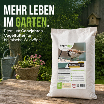 Ein Beutel „TerraUno Premium Wildvogelfutter“ steht auf einem Holztisch im Freien, daneben eine grüne Gießkanne und im Hintergrund blühende Blumen. Der Text hebt dieses hochwertige, schalenlose Ganzjahresfutter als das ideale Vogelfutter für den Garten hervor, mit dem sich Gartenvögel das ganze Jahr über füttern lassen.